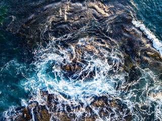 Deep blue water surface. Seaside rocks and foam. Ocean movement top view. Turquoise coastline pattern. Tropical surf texture. Aerial sea waves.