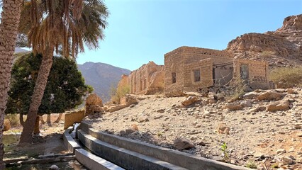Sunlit Irrigation Canal Running Abandoned Adobe Houses And Date Palms, Stone Channel And Walkways Guiding Water Through Arid Settlement, Calm Rural Atmosphere With Strong Lines And Heritage Textures
