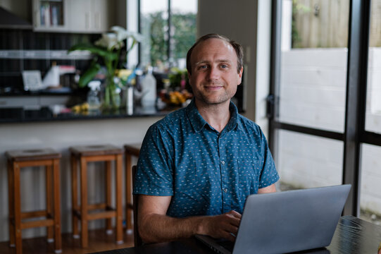 A man sits at a kitchen table, typing on his laptop while smiling