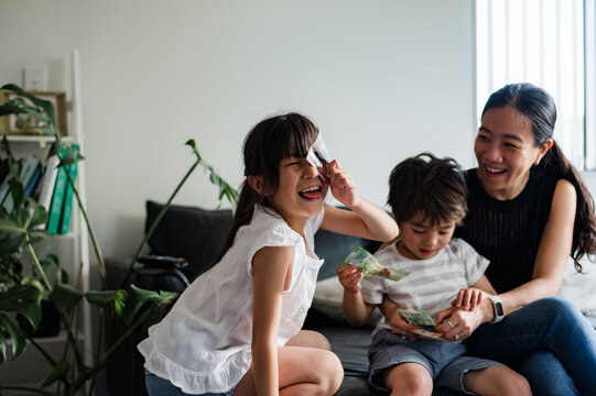Two young children are playing with banknotes while their mother laughs joyfully at home