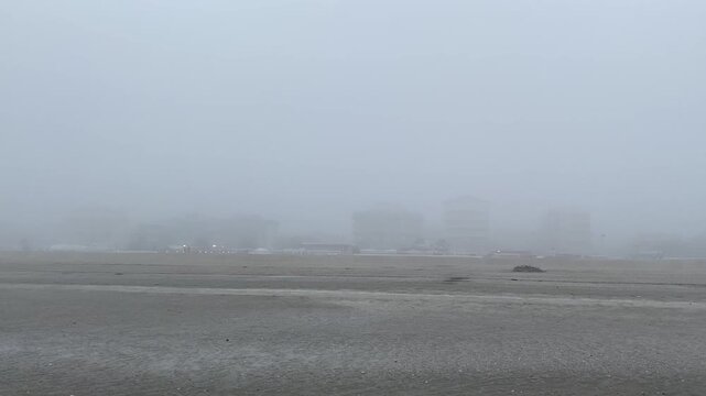 Heavy gray fog obscuring coastal city buildings looming large above the desolate, wet, empty beach in winter. Cervia. Italy