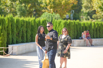 Obraz premium A man and two women are walking in a park. The man is holding a cell phone and the woman is holding a purse. Scene is casual and relaxed