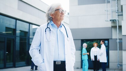 Male senior doctor standing outside hospital building crossing arms while holding confident pose. Experienced Caucasian specialist smiling at camera. Medical workers in background exchanging updates.