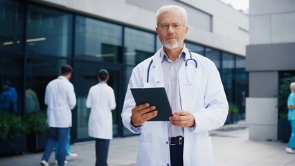 Experienced male doctor standing outside hospital complex. Holding tablet while maintaining focused posture. Experienced Caucasian specialist reviewing patient data. Medical workers in background.