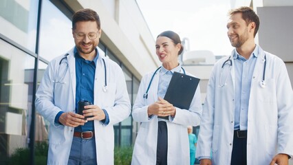 Focused male doctor holding coffee and walking with smiling colleagues outside clinic. Confident female carrying clipboard and discussing medical tasks. Male companion sharing ideas while moving along