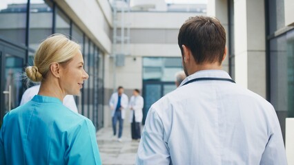 Smiling female nurse walking beside focused male doctor outside clinic. Confident pair discussing duties while moving along pathway. Professional team sharing thoughts during relaxed moment.