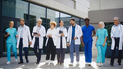 Medical team walking toward camera outside clinic showing coordinated movement. Confident doctors and nurses carrying tablets and reviewing duties while moving together. Preparing for upcoming tasks.