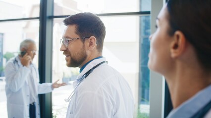 Close camera view from behind two focused doctors walking through bright clinic hallway. Professionals talking casually while moving toward duties. Team preparing for busy day together.