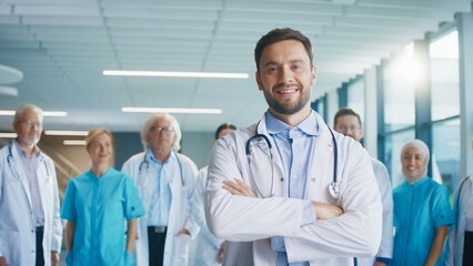Smiling male doctor standing in spacy corridor and crossing arms. Friendly medical professional greeting supportive team behind him. Showing calm confidence. Enjoying working at hospital.