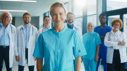 Beautiful female nurse standing close to camera in bright hospital corridor. Smiling confidently. Colleagues standing behind in blurred background supporting teamwork during active clinical routine.