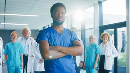 Confident male medical worker crossing arms while standing in bright corridor. Focused nurse presenting strength while group of doctors supporting him in background. Preparing for challenging duties.