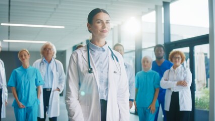 Confident female doctor standing at front of camera. Focused medical specialist crossing arms while team gathering behind her. Preparing for upcoming responsibilities in busy hospital setting.