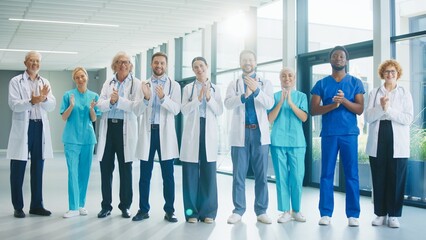 Medical team standing in bright hospital hall clapping toward camera. Smiling proudly. Showing appreciation for teamwork and celebrating achievements while enjoying positive moment.