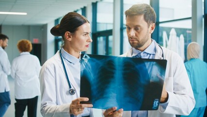 Two doctors standing in bright hospital corridor holding chest X-ray. Studying image carefully. Discussing findings and evaluating condition while planning next diagnostic steps together.