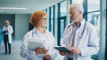 Two doctors standing in bright hospital corridor holding documents and tablet. Talking about patient diagnosis. Reviewing details and coordinating treatment plan while preparing for next steps.