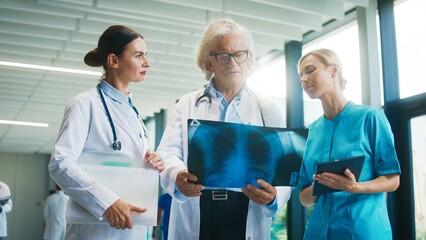 Three medical professionals reviewing chest X-ray together in bright hospital corridor. Senior doctor holding image while younger colleagues discuss. Team collaborating and evaluating condition.