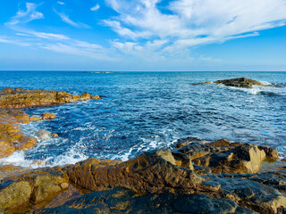 Waves crash on rocks along the coastline under a blue sky with clouds near the ocean during the day