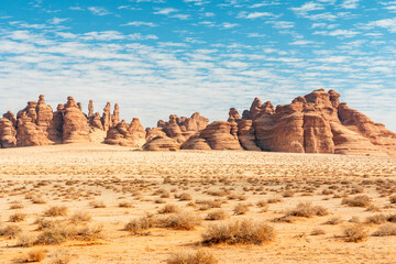 Desert Hegra, Saudi Arabia landscape with eroded rock formations, sparse scrub, dramatic sunlit open terrain, bright blue sky