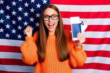 Happy woman in orange knit sweater poses with phones in front of American flag for celebration...