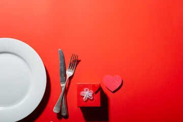 Empty plate with cutlery by heart shape and valentine present box on red background with copy space
