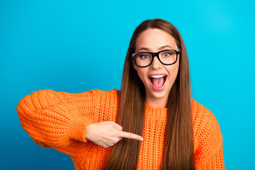 Young woman in orange sweater smiling excitedly points at camera in bright blue studio setting...