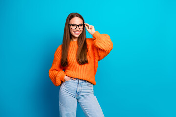 Young woman in orange knit sweater smiles while adjusting glasses against bright blue background