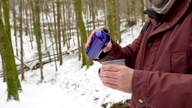 Middle aged man with a backpack taking a break to pour and drink a warm beverage from a flask while hiking through a beautiful snowy forest during a cold winter day, enjoying nature