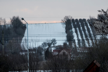 Ośnieżone wzg&oacute;rza z polami i sadami w wiejskim krajobrazie pod zachmurzonym, zimowym niebem. Rzędy bezlistnych drzew wzdłuż p&oacute;l tworzą wzory na śniegu. Na pierwszym planie linie energetyczne 
