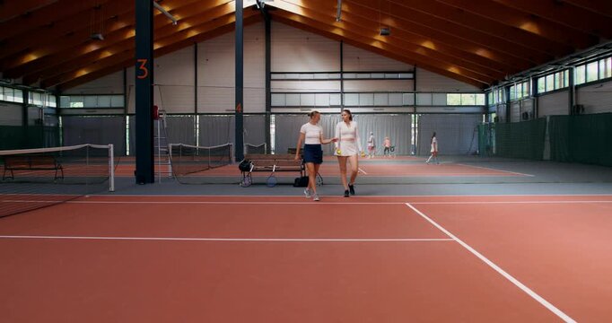 Female tennis players take balls and go out on the court to train. Two female tennis players do warm-up exercises, then begin training.