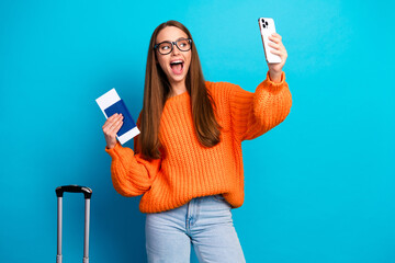 Young woman in orange knit sweater takes a selfie with phone and passport against blue background