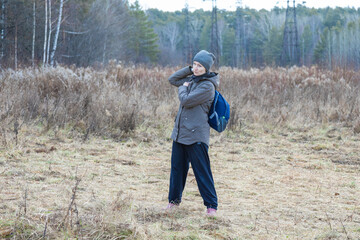 Woman tourist with backpack stands in a rural field with dry grass, adjusting her backpack while looking ahead. Outdoor adventure and hiking in nature.