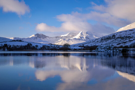 A Winter landscape of snow-covered mountains, highlighted by the sun and reflected in the still waters of  a lake. Llyn Mymbyr, Capel Curig, Snowdonia, Wales,