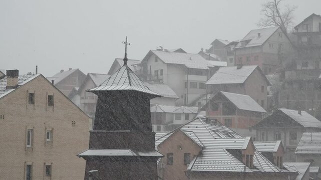 Snow falls steadily over St. Mark Orthodox Church and surrounding homes in Uzice, Serbia. A quiet winter town with tiled rooftops, church tower, and overcast sky creates a calm urban atmosphere