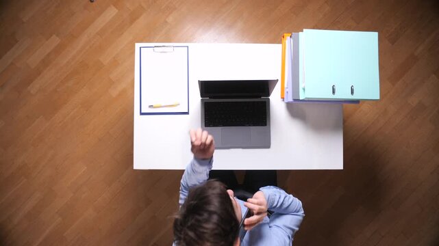 Business professional multitasking at a white desk, on a phone call while managing laptop, clipboard and file folders in a top view office scene conveying urgency and productivity
