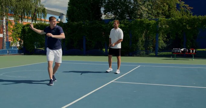 A guy practices his swing technique on the tennis court. Two male tennis players train on the court with rackets in hand.