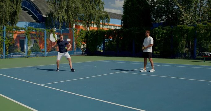 Two guys practice on the tennis court. Two male tennis players train on the court with rackets in hand.