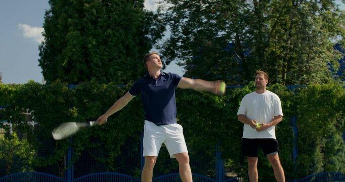 A player picks up the ball and hits it with a racket.. Two male tennis players train on the court with rackets in hand.