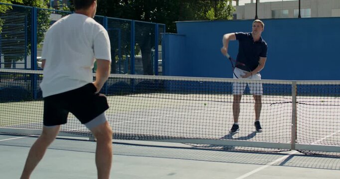 Two guys practice passing the ball over a tennis net.. Two male tennis players train on the court with rackets in hand.