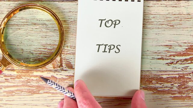 Hands turn pages revealing top tips written on a notepad, next to a golden magnifying glass and patterned pen, all resting on a weathered wooden surface