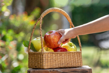 Hand pick up apple from a wicker basket filled with fresh red and green apples outdoors in natural sunlight, symbolizing harvest, organic fruit, healthy eating, and farm to table lifestyle.