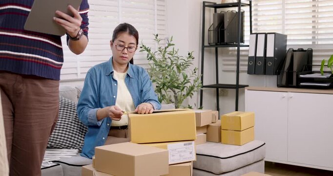 Asian woman online business owner sealing parcel box, checking shipping label placement, preparing online shopping parcel box for delivery during daily e-commerce workflow at home