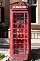 Fototapeta premium Weathered red telephone booth standing in front of historic limestone buildings, symbol of traditional public communication and urban heritage. Malta, December 30, 2025.