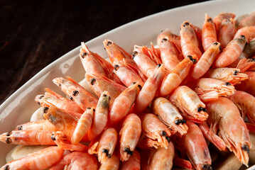A close-up horizontal shot of cooked shrimp with their heads on, piled high on a white serving platter with a dark, rustic background.