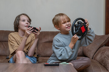 Two brothers playing video games on a couch at home. Kids with smartphone and toy wheel