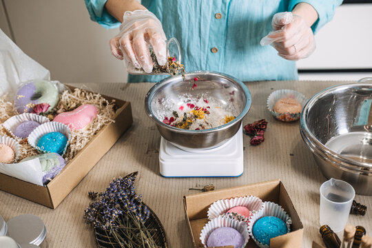 Hands in gloves pour botanical mixture from glass beaker into metal bowl on digital scale. Small-batch production, artisan cosmetics, indie beauty brand, cosmetic formulation.