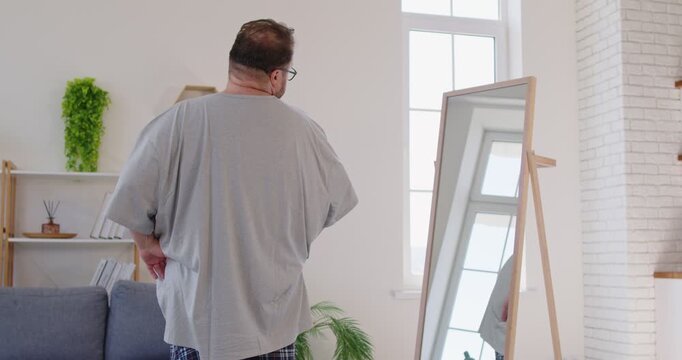 Obese confident man looking at mirror reflection at home. Happy proud male person with obesity wearing oversize tee shirt, body positive guy holding and stroking big belly with pride and posing