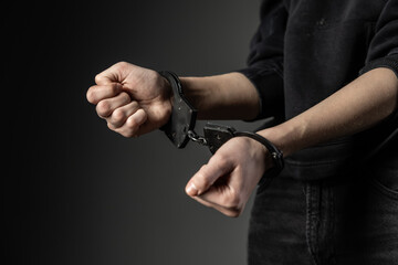 A close-up of a person&rsquo;s wrists locked in black handcuffs, set against a dark background. The tense pose and cropped composition emphasize confinement and emotion