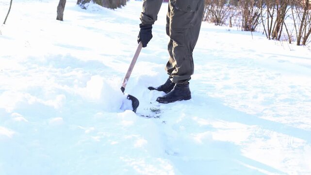 A person shoveling snow in a winter landscape. The individual wears dark pants and boots, working to clear a path in the white snow.