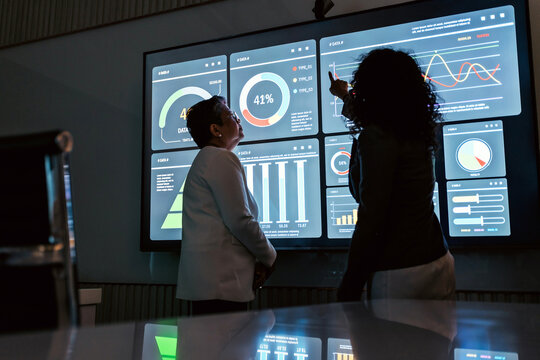 Two professionals analyze data on a large touchscreen, pointing at colorful dashboards and graphs in a dark office, illustrating analytics, teamwork, and decision making. - Powered by Adobe