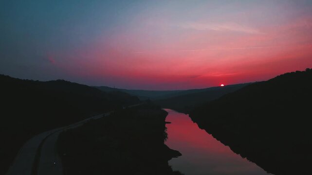 Cinematic slow upward shot of a small river at dusk, reddish sunset reflections on calm water, dark serene mood with a parallel highway adding subtle urban contrast.